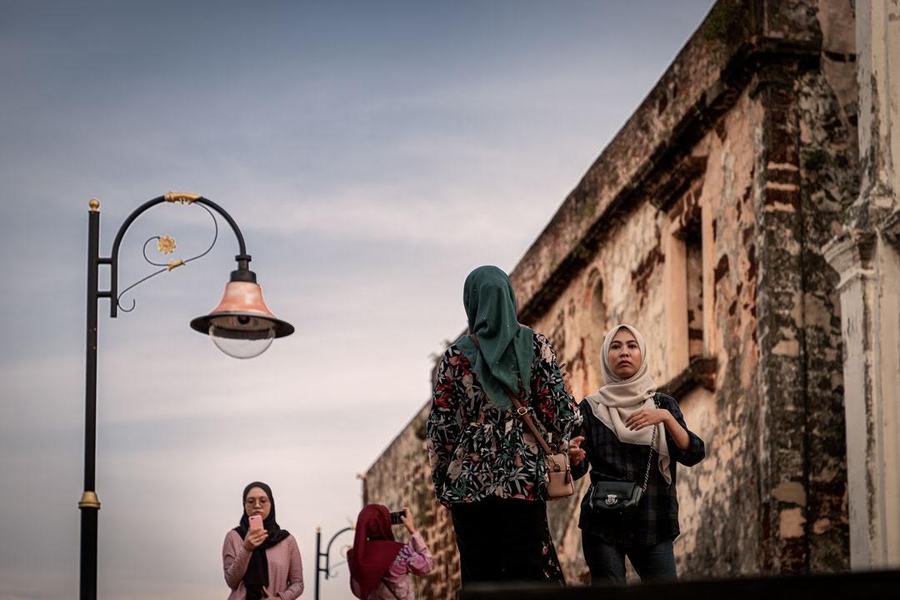 Two women in headscarves pause on a street beside a weathered old fort wall, an ornate street lamp overhead.